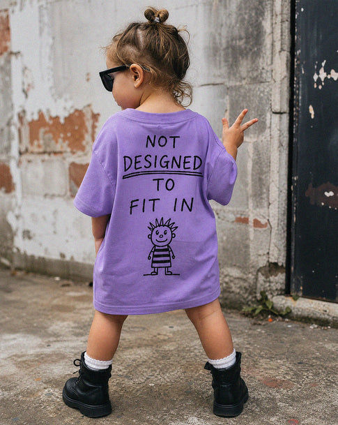 Child wearing a purple shirt with Not designed to fit in text and a graphic, standing against a textured wall.