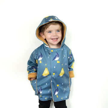 Little boy wearing a lottie & lysh pigeon puddles splasher jacket. The jacket is blue, with pigeons wearing yellow rain coats, hats and wellies. The fabric also features umbrellas and raindrops.