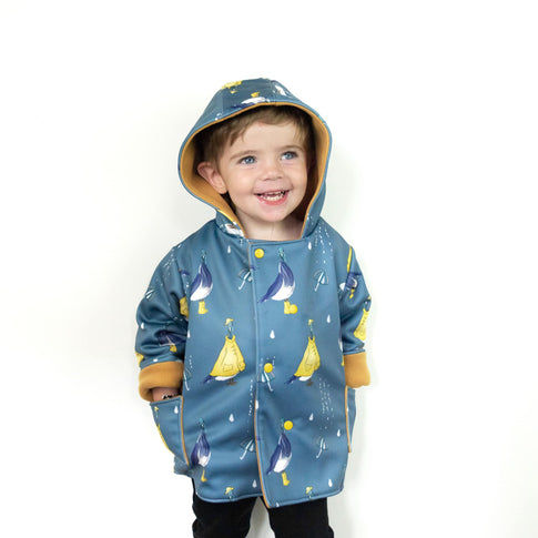 Little boy wearing a lottie & lysh pigeon puddles splasher jacket. The jacket is blue, with pigeons wearing yellow rain coats, hats and wellies. The fabric also features umbrellas and raindrops.