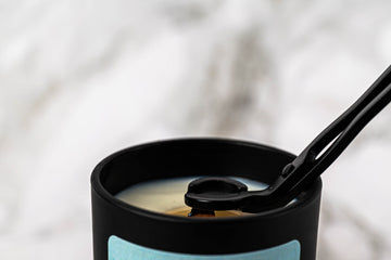 A close-up of a black candle with a wick trimmer cutting the candle wick. The background is softly blurred in white and gray tones.