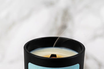 A close-up of a black candle jar with a recently extinguished wick, releasing a thin trail of smoke against a blurred, light background.
