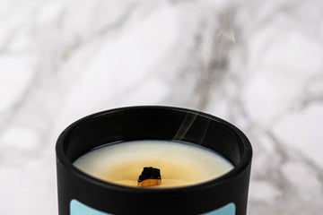 A black candle jar with a wooden wick sits on a marble surface. The candle is extinguished, and a thin trail of smoke rises from the charred wick.