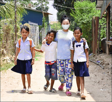Four children in school uniforms and backpacks stand together on a dirt path, smiling. One child wears a mask. Trees and houses are visible in the background on a sunny day.