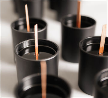 Several black matte candle jars with wooden wicks are arranged closely together on a light surface, seen from above and at an angle. The candles appear unlit and unused.