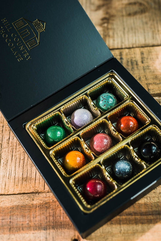An open black box with gold lining displays nine assorted Hill Country Chocolate Signature Artisanal Bonbons—a beautiful, handmade collection that’s sure to delight any chocolate lover.