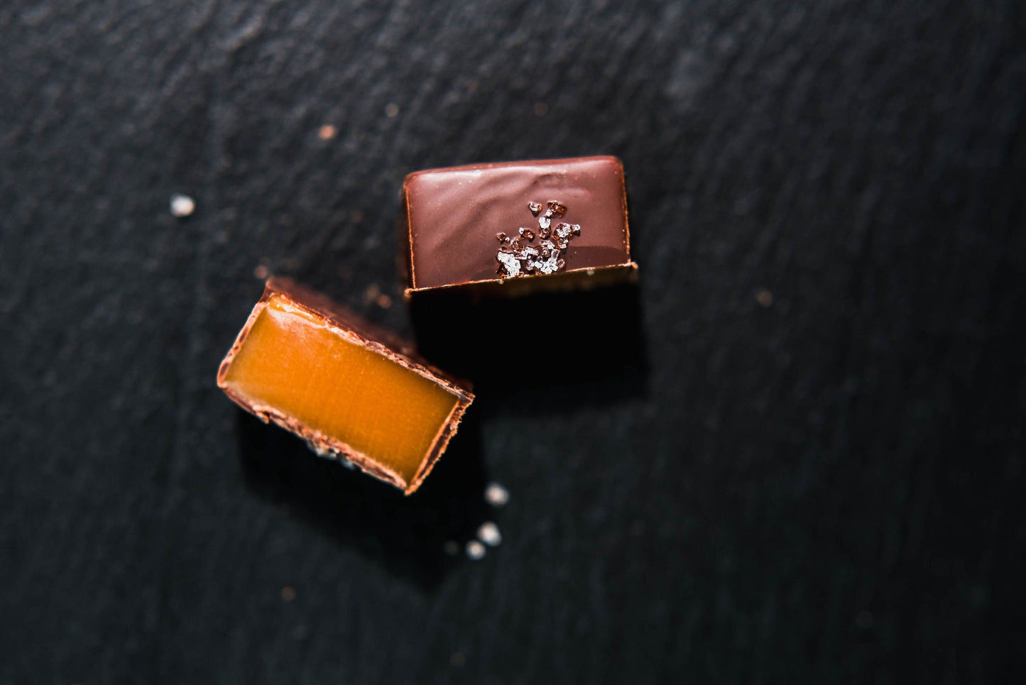 Two caramels from the Hill Country Chocolate 15 piece Mixed Caramels box, one cut open to reveal its rich caramel center, rest on a dark slate with sea salt—a decadent treat inspired by Texas Hill Country chocolates.