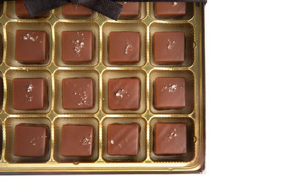 A gold tray of Hill Country Chocolate's 15 piece Mixed Caramels, inspired by Texas flavors and topped with coarse salt, is partially covered by a black ribbon and set against a white background.