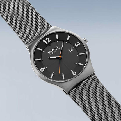 Herrenuhr mit schwarzem Zifferblatt, orange Zeiger, runde Lünette und Metall-Meshband