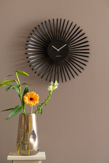 Svart väggklocka på väggen med radiella ränder, bredvid blomsterbukett i vas