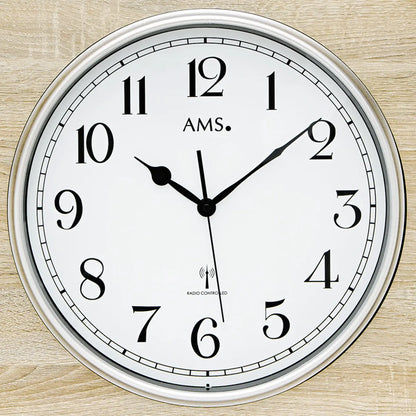 Nahaufnahme: Funk-Wanduhr mit AMS-Logo, weißem Zifferblatt, schwarzen Zeigern und Funkhinweis