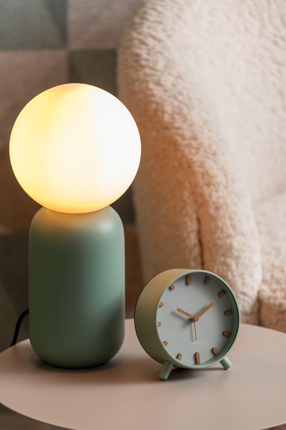 Grågrön väckarklocka på bord bredvid sfärisk lampa med varmt ljus