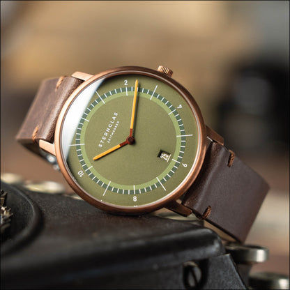 Herrklocka i snedbild: rund boett, grön urtavla, orange visare, läderarmband
