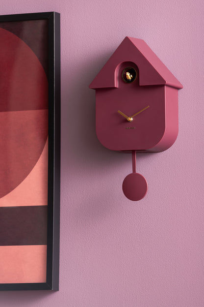 Väggklocka i fågelhusdesign, mörkrött utförande med visare och runt pendel på väggen.