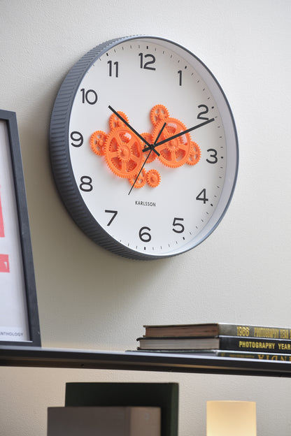 Väggklocka på väggen med grå ram och orange synligt urverk