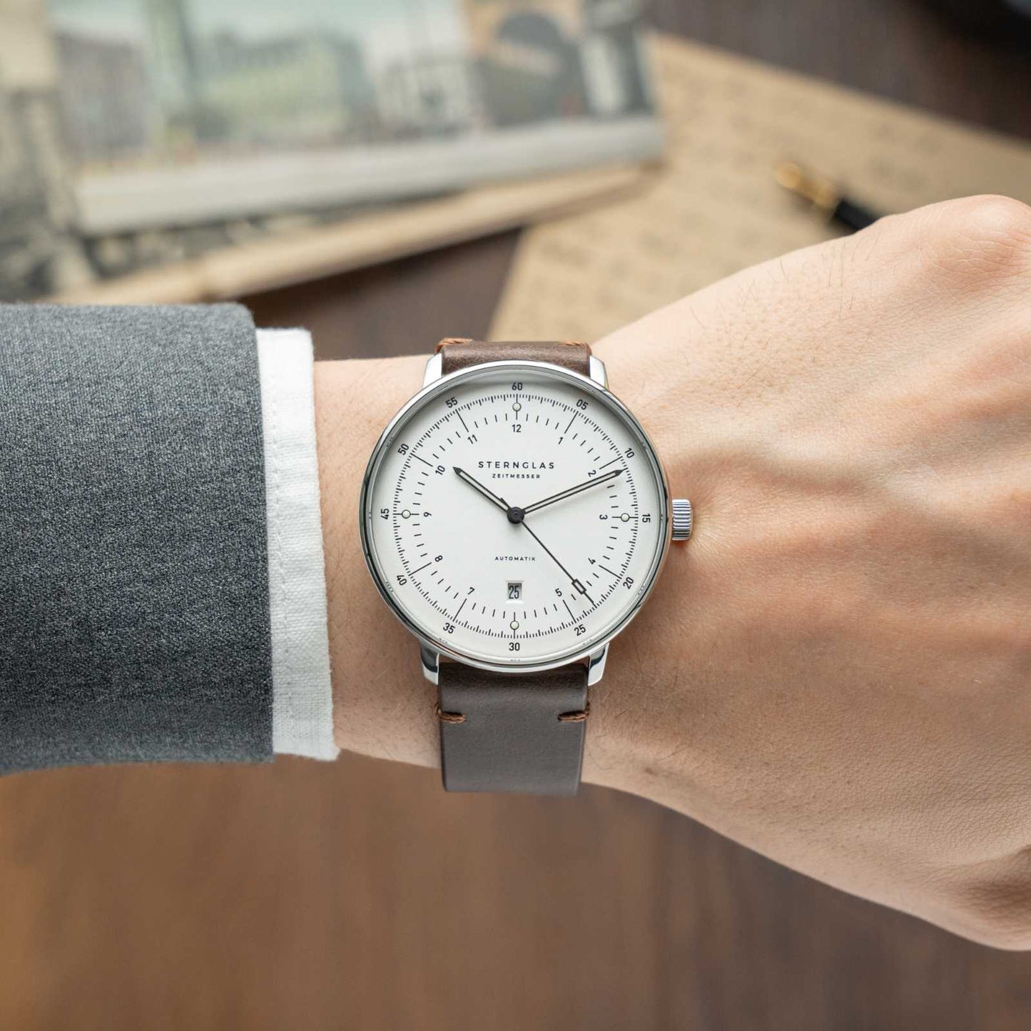 Herrklocka på handleden: brunt läderarmband, silverfärgat hölje, vit urtavla med index.