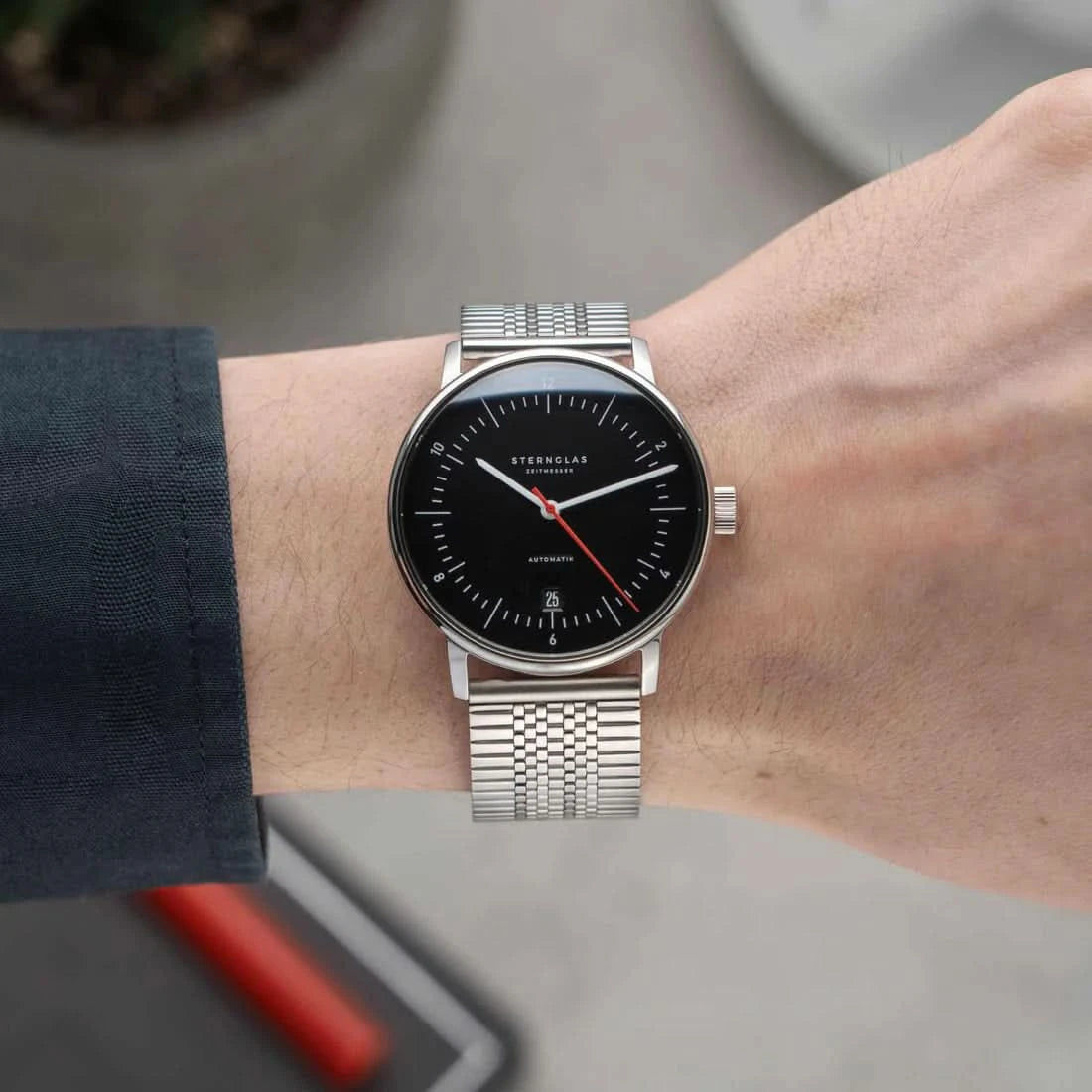Herrklocka på handleden: svart urtavla, röda visare, silverfärgat mesh-armband
