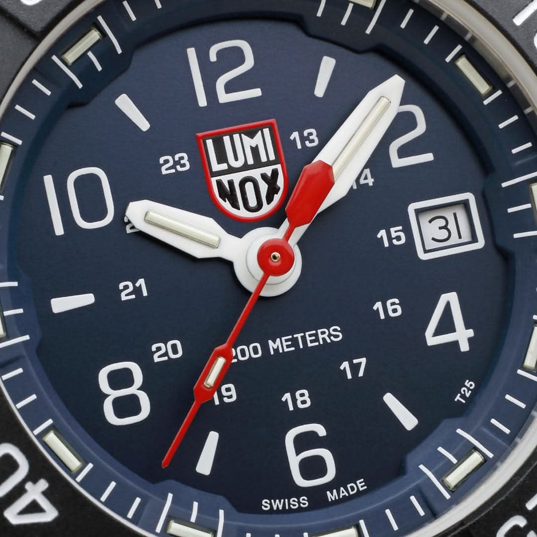 "Närbild av urtavla: vita index, röd sekundvisarspets, LUMINOX-logotyp och datumfönster."