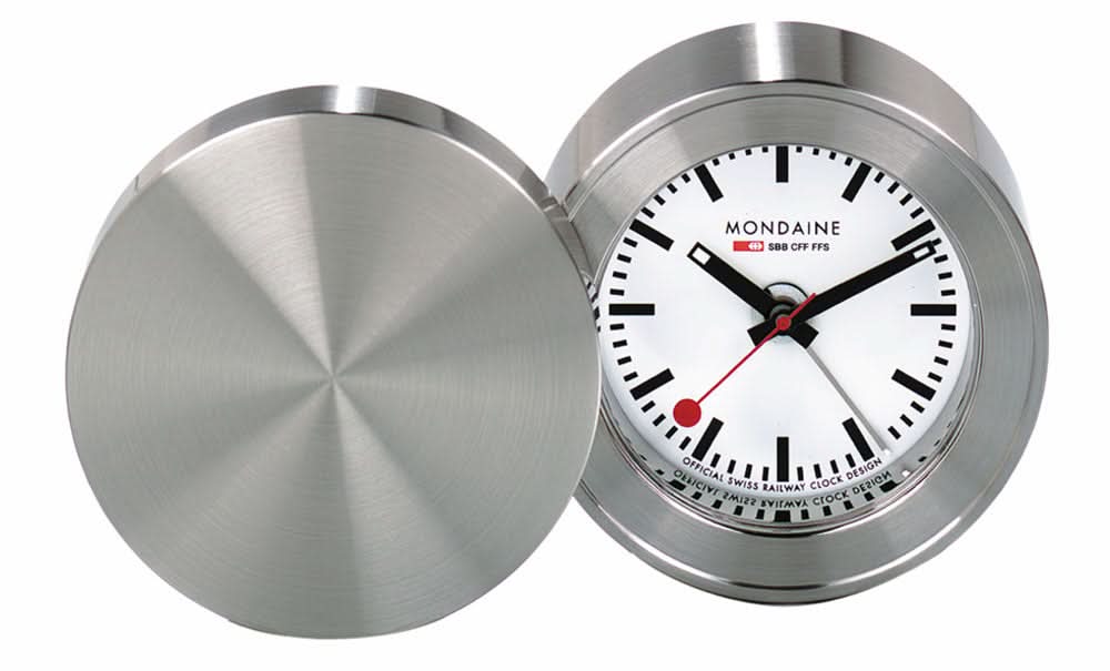 Mondaine reseväckarklocka med öppet silverfärgat lock, runt metallhölje och vit urtavla