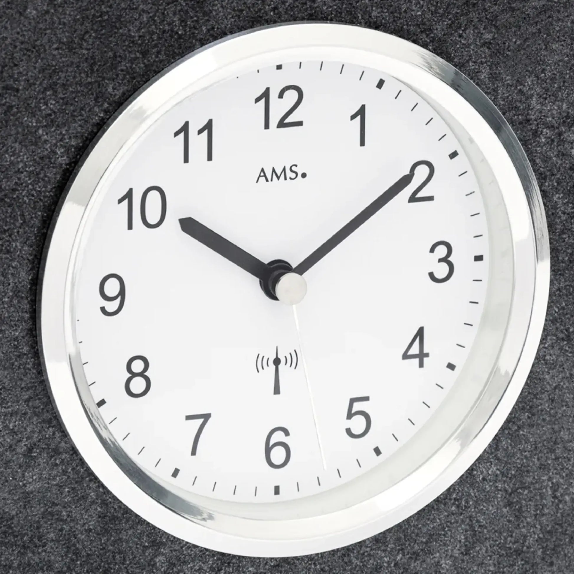 Nahaufnahme: rundes Zifferblatt mit AMS-Schrift und Funkwellen-Symbol, schwarze Zeiger.