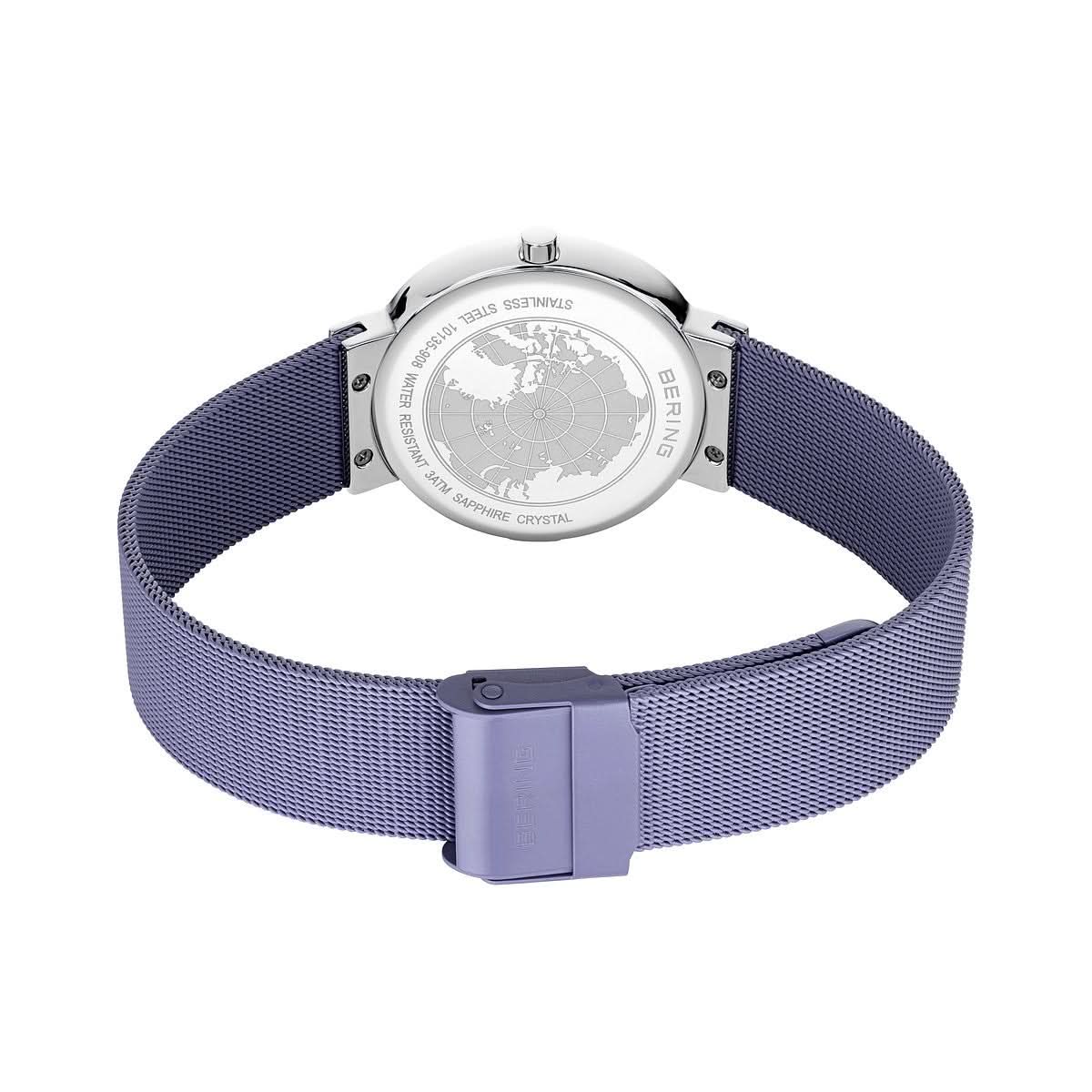 Reloj de mujer con correa Milanesa morada y cierre; fondo de acero inoxidable visible.