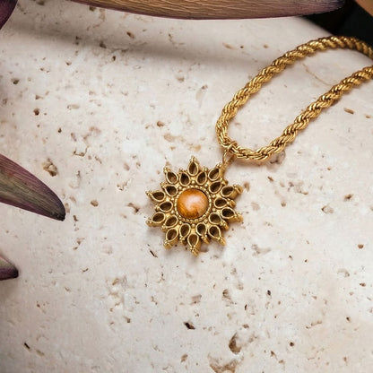 Colgante de sol en cadena dorada con cabujón marrón, sobre superficie de piedra clara