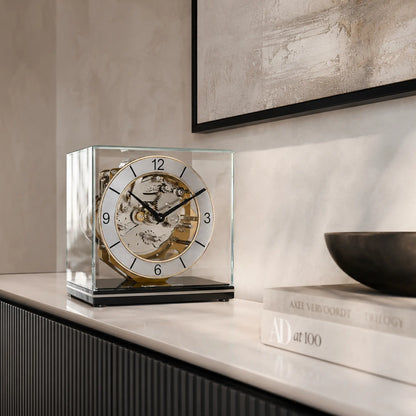 Reloj de mesa en caja de cristal con mecanismo mecánico visible, esfera redonda y números romanos