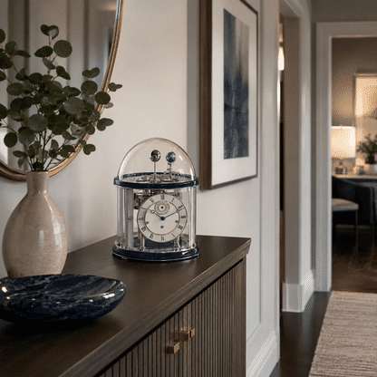 Reloj de mesa con campana de cristal sobre consola de madera, esfera plateada, base azul