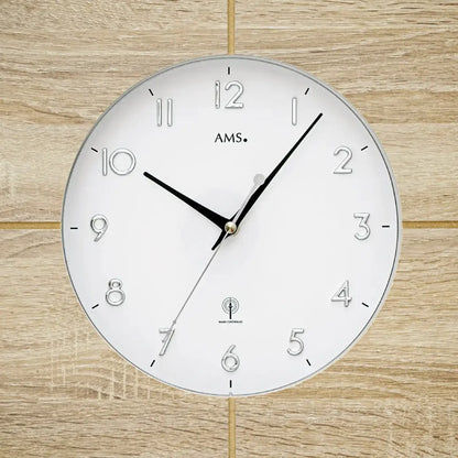 Nahaufnahme Wanduhr AMS: weißes Ziffernblatt mit Zahlen 1–12 und schwarzen Zeigern