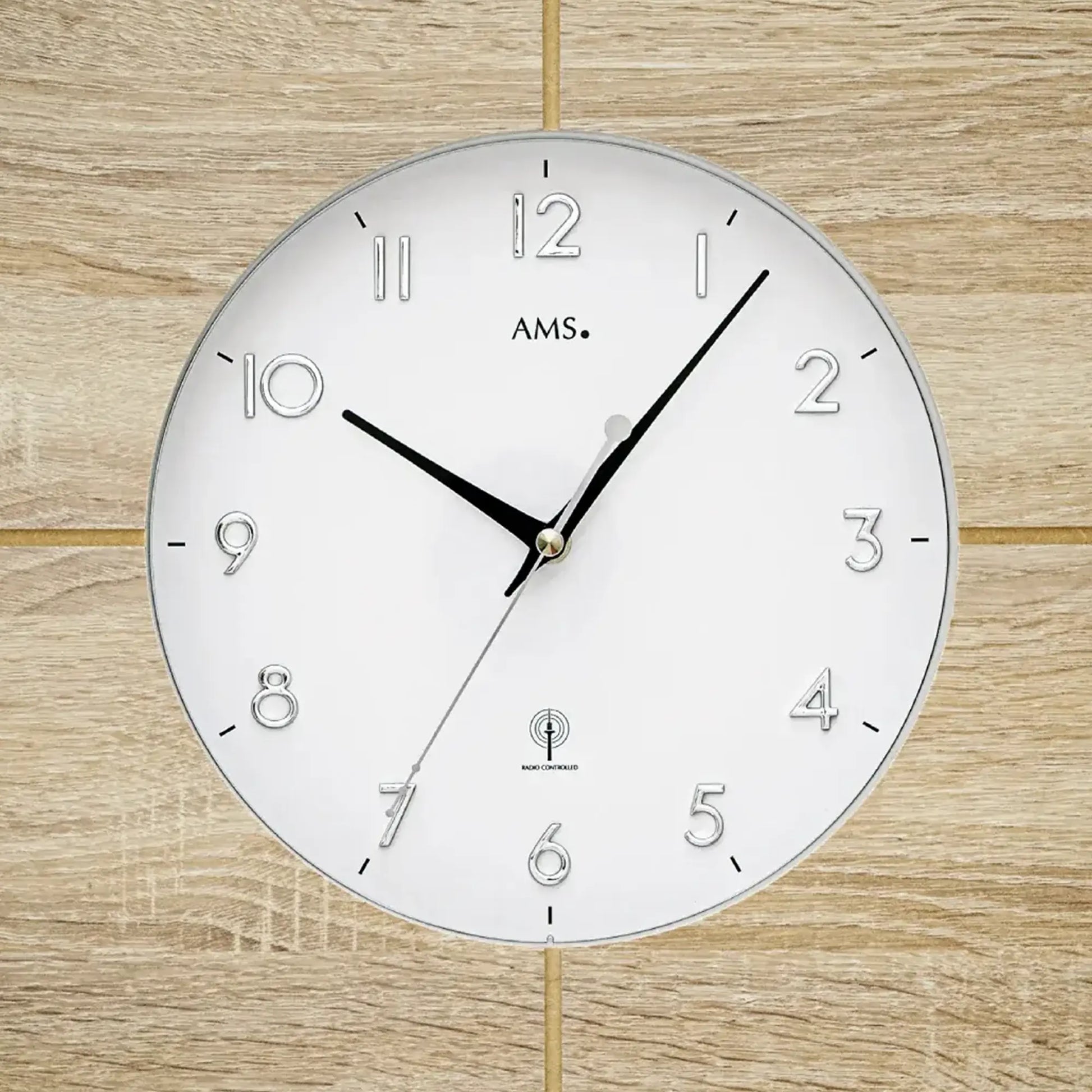Nahaufnahme Wanduhr AMS: weißes Ziffernblatt mit Zahlen 1–12 und schwarzen Zeigern