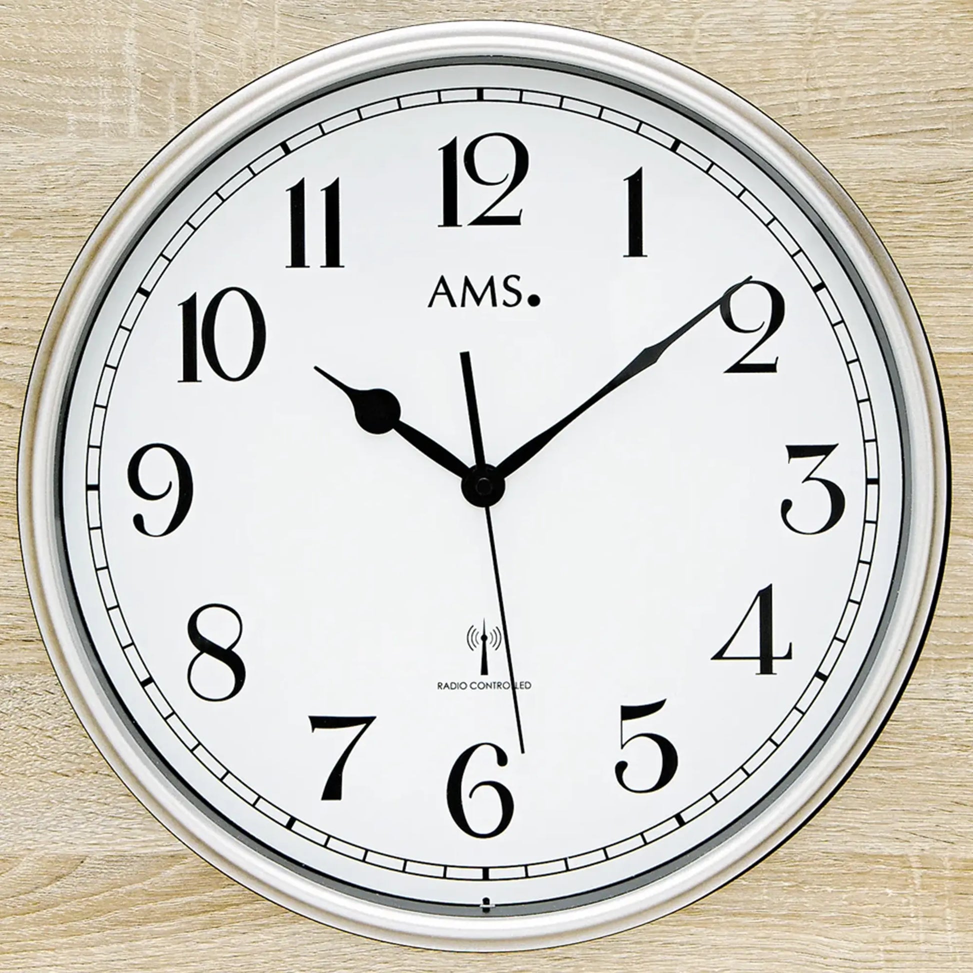 Nahaufnahme: Funk-Wanduhr mit AMS-Logo, weißem Zifferblatt, schwarzen Zeigern und Funkhinweis