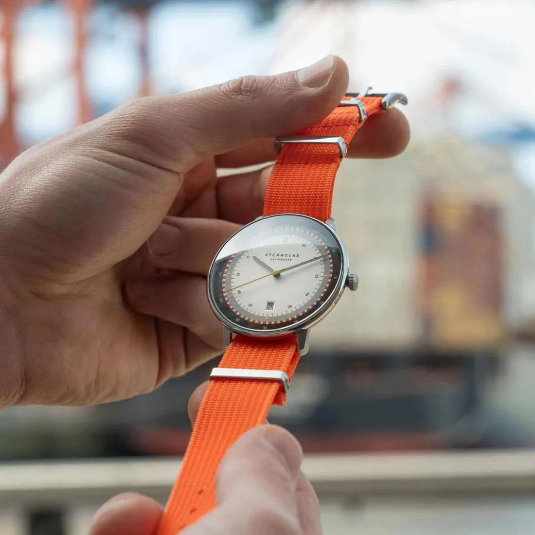 Herrenuhr am orangefarbenen Textilarmband gehalten, rundes weißes Zifferblatt
