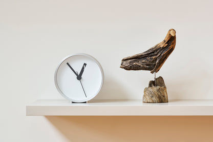 Weiße runde Wanduhr auf Regalbrett mit schwarzen Zeigern, daneben hölzerne Deko.