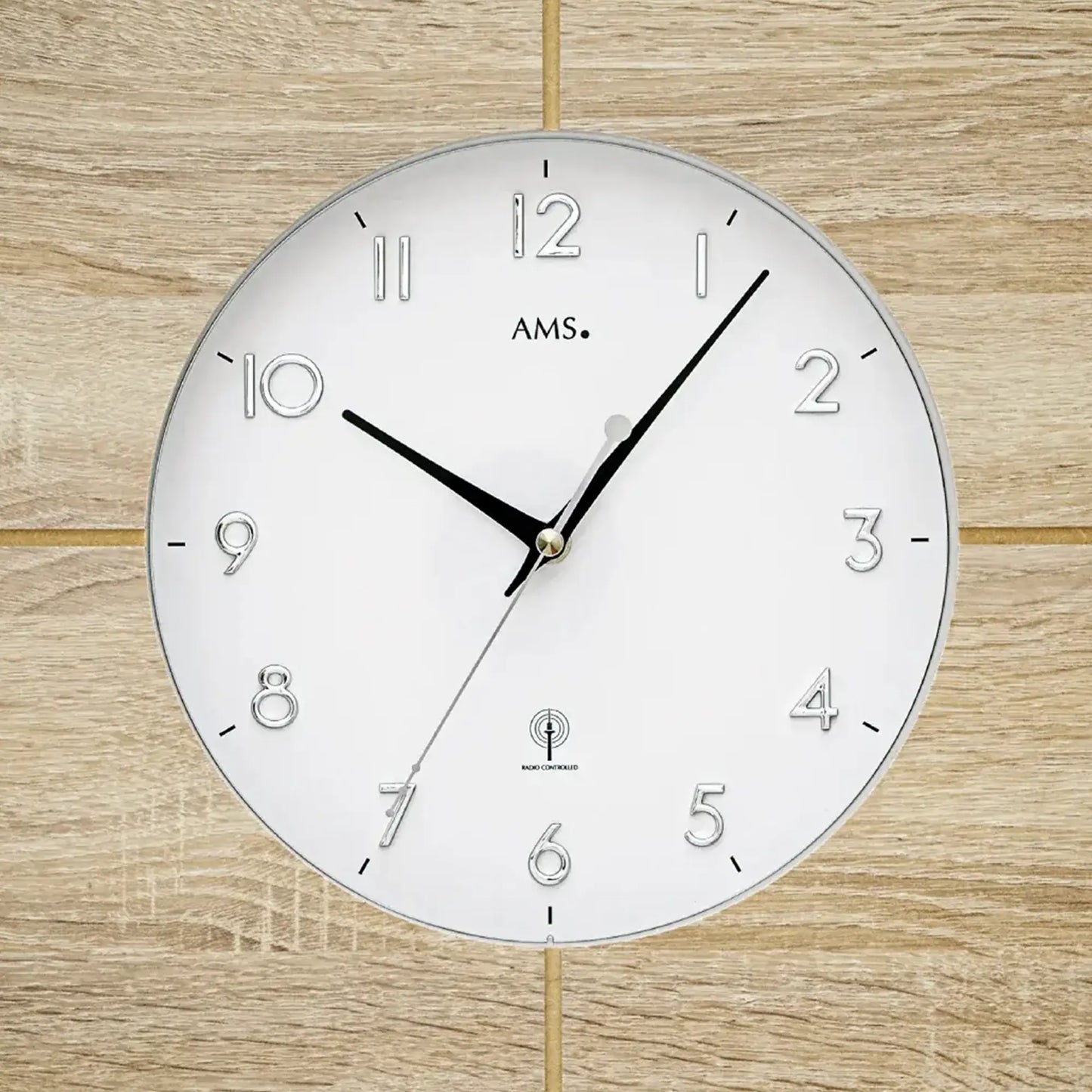 Nahaufnahme Wanduhr AMS: weißes Ziffernblatt mit Zahlen 1–12 und schwarzen Zeigern
