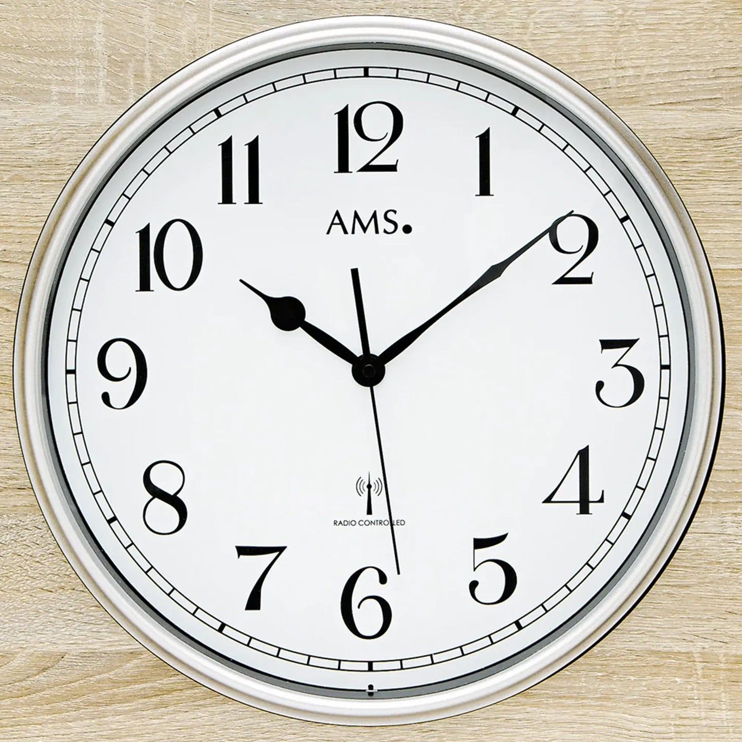 Nahaufnahme: Funk-Wanduhr mit AMS-Logo, weißem Zifferblatt, schwarzen Zeigern und Funkhinweis