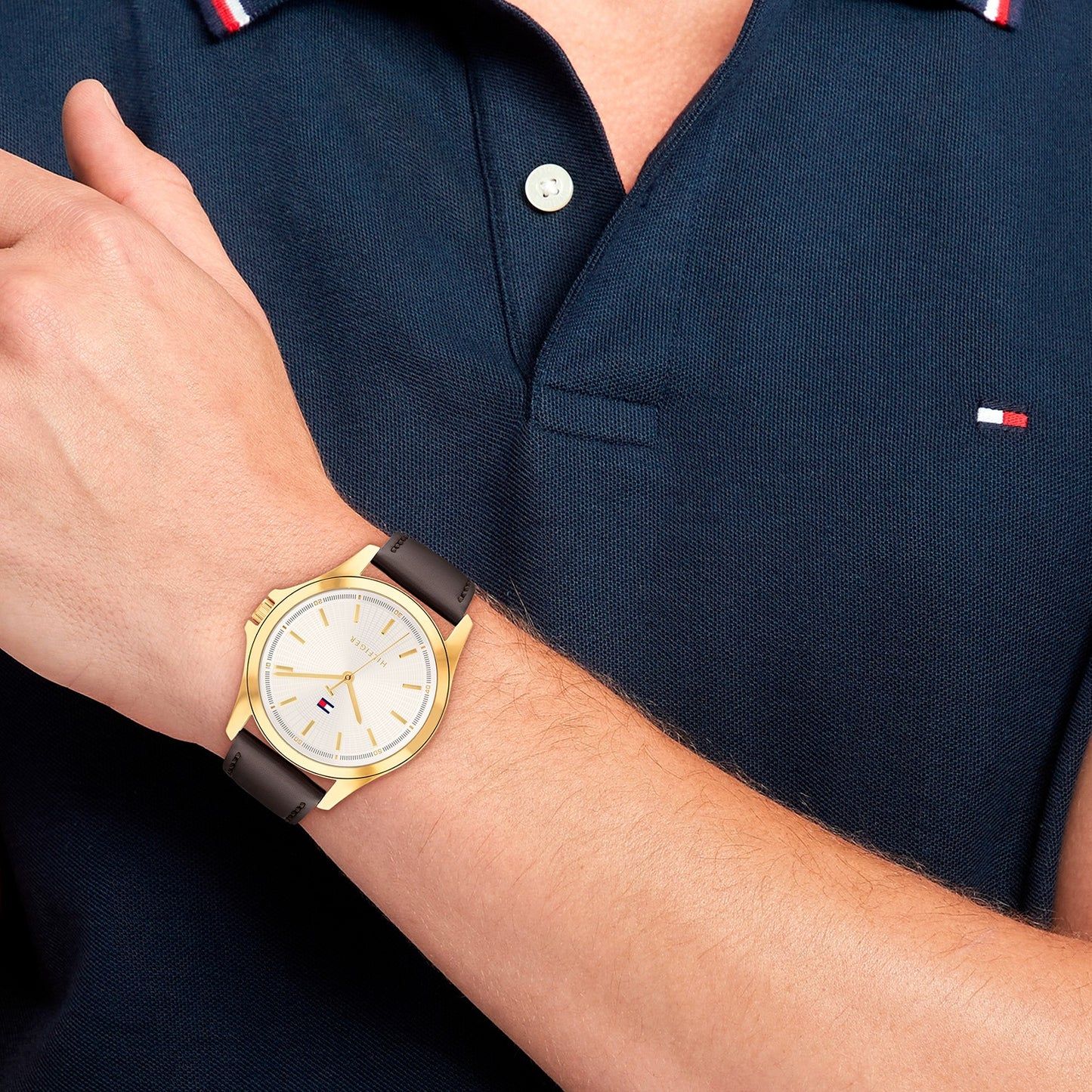 Tommy Hilfiger Herren-Armbanduhr am Handgelenk: weißes Zifferblatt, dunkles Lederband, goldene Lünette.