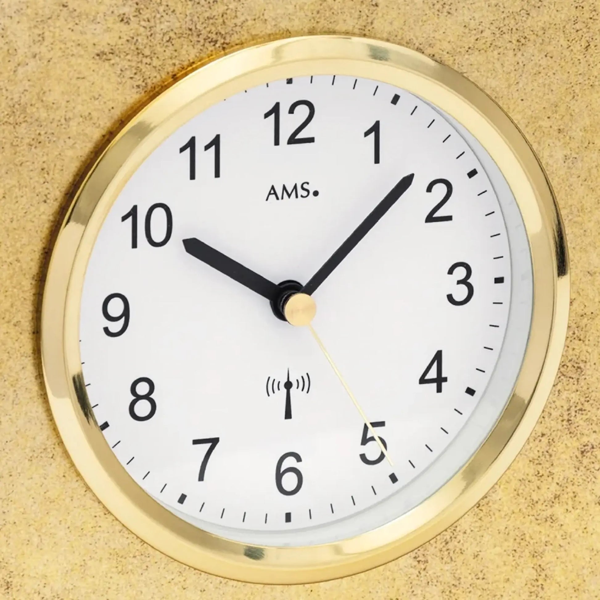 Nahaufnahme Tischuhr: weißes Ziffernblatt mit AMS-Logo, goldener Rahmen, Funk-Symbol